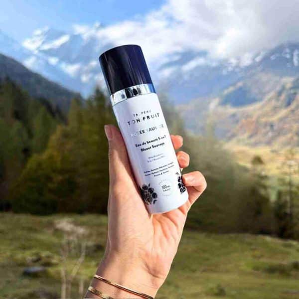 Eau de Beauté 5-en-1 Rosée Sauvage au Bleuet Sauvage tenue en main devant un paysage de montagne