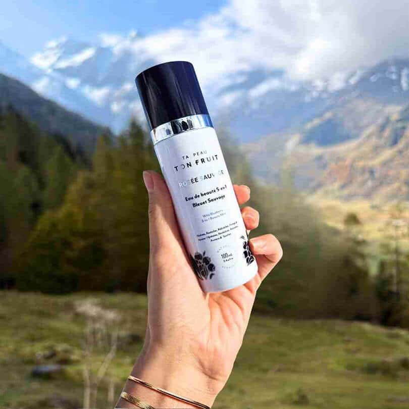 Eau de Beauté 5-en-1 Rosée Sauvage au Bleuet Sauvage tenue en main devant un paysage de montagne