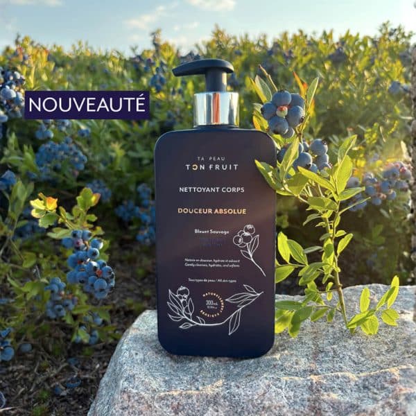 Bouteille de Nettoyant Corps Douceur Absolue au Bleuet Sauvage tenue en main devant des buissons de bleuets