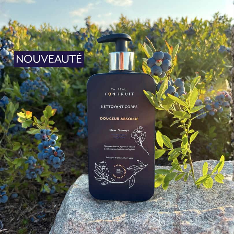 Bouteille de Nettoyant Corps Douceur Absolue au Bleuet Sauvage tenue en main devant des buissons de bleuets